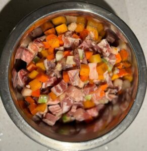 Металлическая миска сверху, доверху наполненная нарезанным кубиками мясом и овощами: морковь, кабачок, кусочки сельдерея вперемешку с кусками мяса с прослойками жира