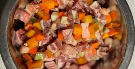 Металлическая миска сверху, доверху наполненная нарезанным кубиками мясом и овощами: морковь, кабачок, кусочки сельдерея вперемешку с кусками мяса с прослойками жира