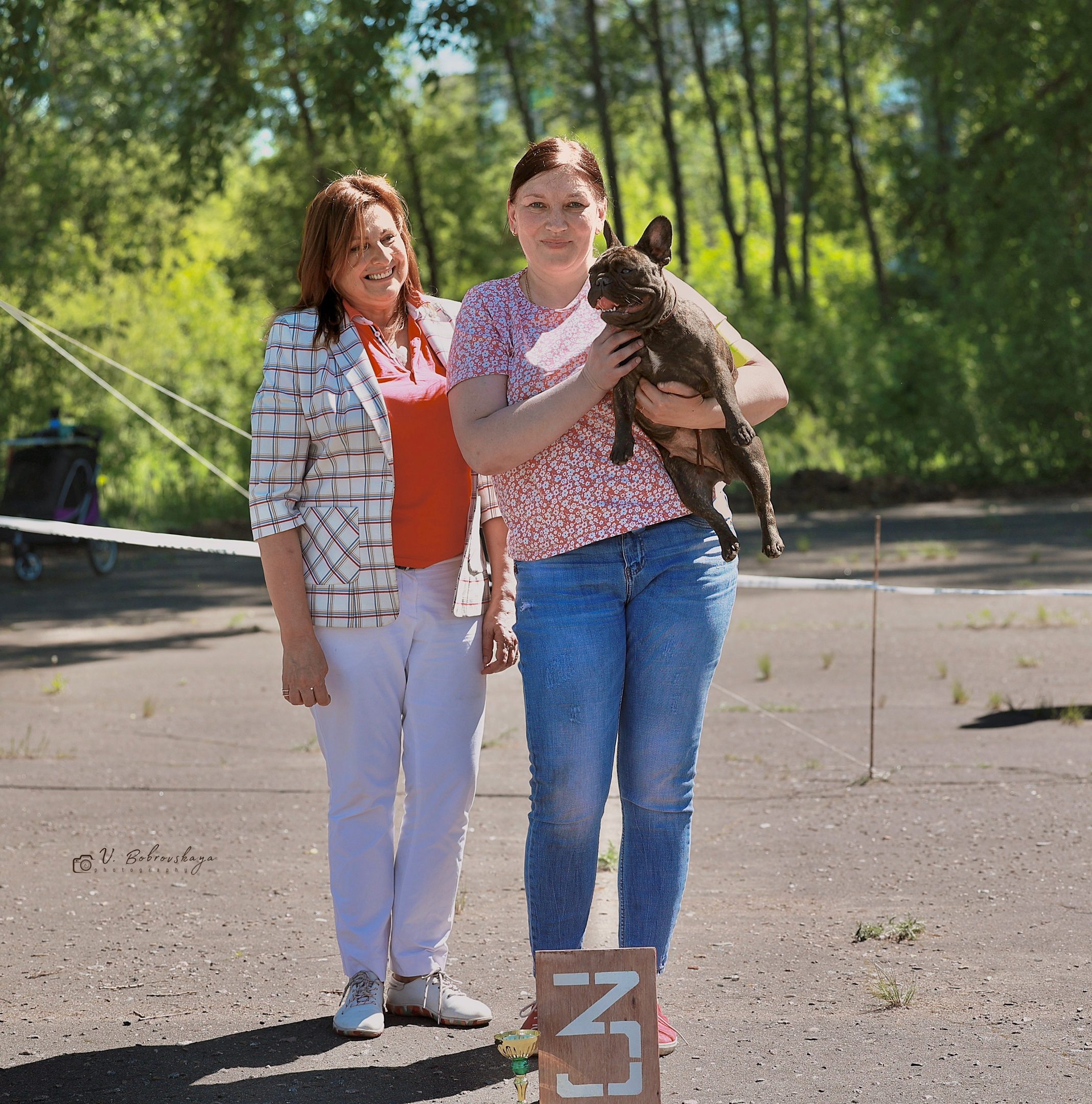 Иштар юниором на руках у владелицы рядом с судьёй на площадке в лесу, у ног табличка третьего места
