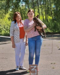 Иштар юниором на руках у владелицы рядом с судьёй на площадке в лесу, у ног табличка третьего места